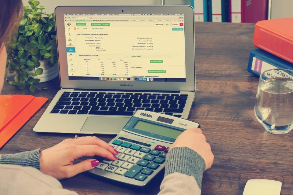 Hands using a calculator beside a laptop showing an accounting ledger